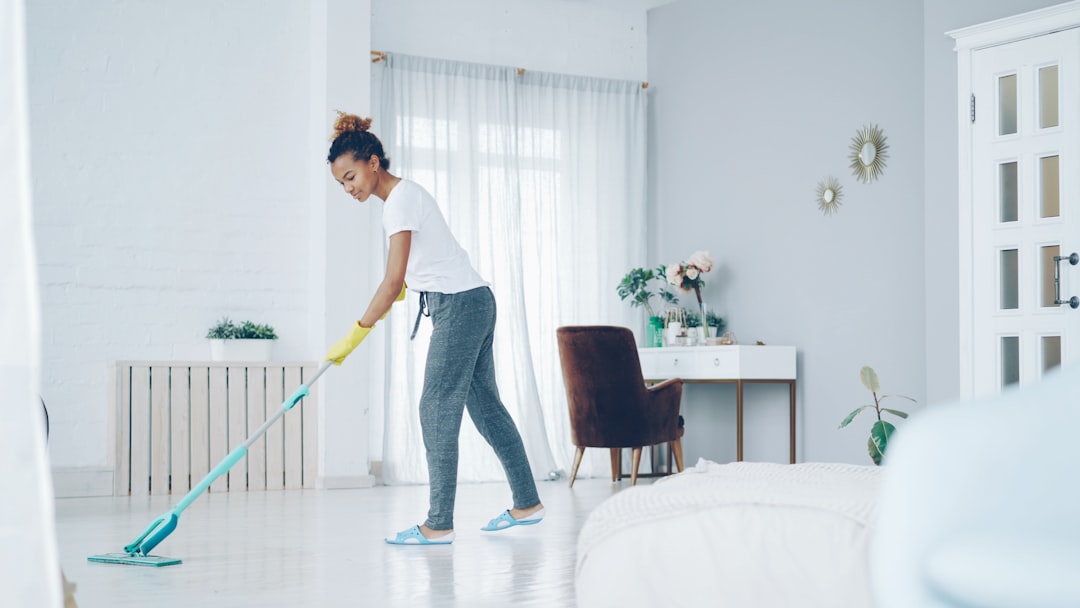 A pretty African American girl is mopping the floor at home with a flat plastic mop, wearing gloves and casual at-home clothes. Modern interiors, cleanliness, and young people concept.